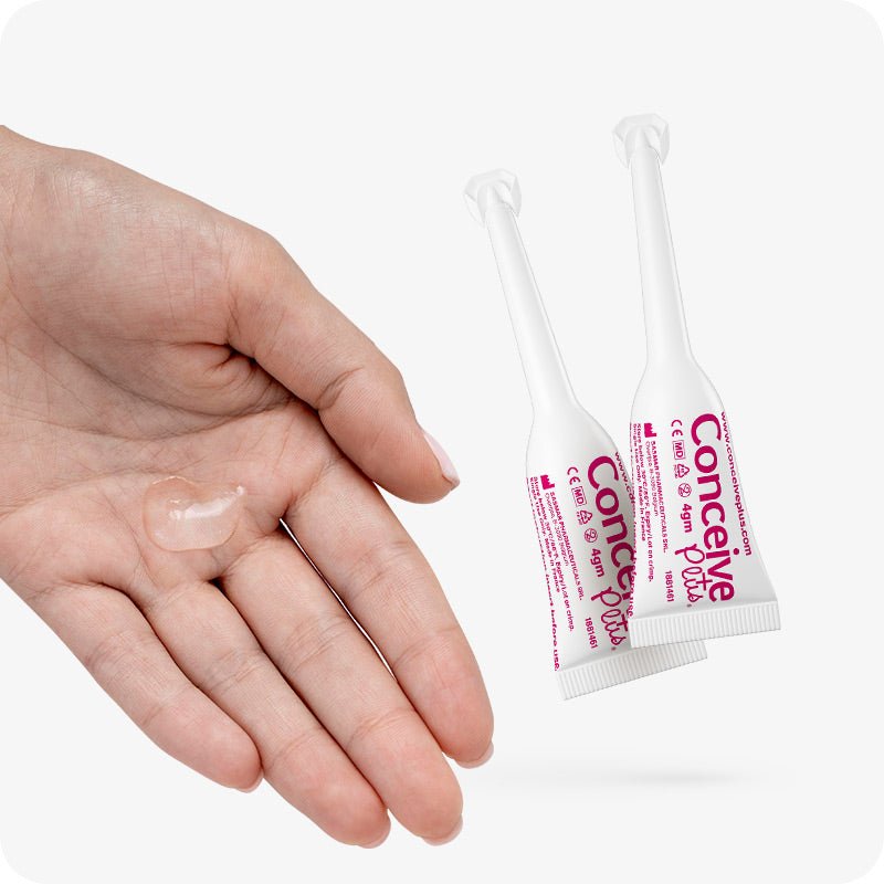 Image of two Conceive Plus fertility lubricant, pre-filled applicators 4g and a hand with small quantity of lube on palm of a female hand on a white background. Applicators are on right and hand on left. White applicators has pink branding with information about website url, manufacturer details, and storage directions.