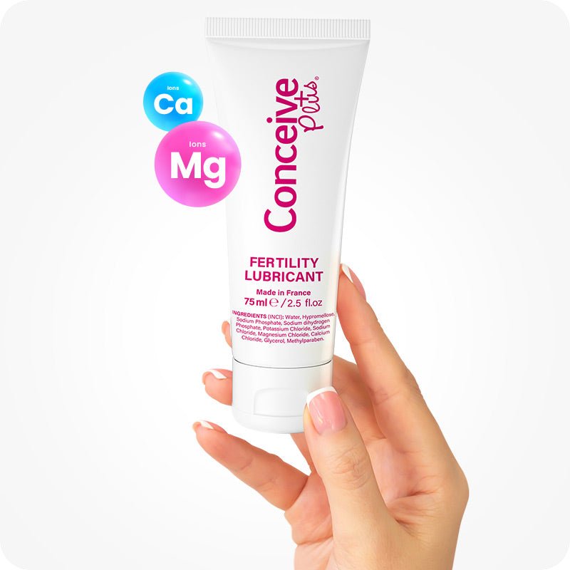 Image of Conceive Plus fertility lubricant tube 75ml being held by a female hand on a white background. White tube with pink branding and text highlights information about quantity of lube, ingredients, and made in France text. Two Calcium and Magnesium ion bubbles are floating in the frame.