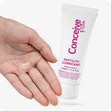 Image of Conceive Plus fertility lubricant tube 75ml and a hand with small clear lube on its palm on a white background. White tube has pink text and branding with highlights about lube quantity, ingredients, and made in France text.
